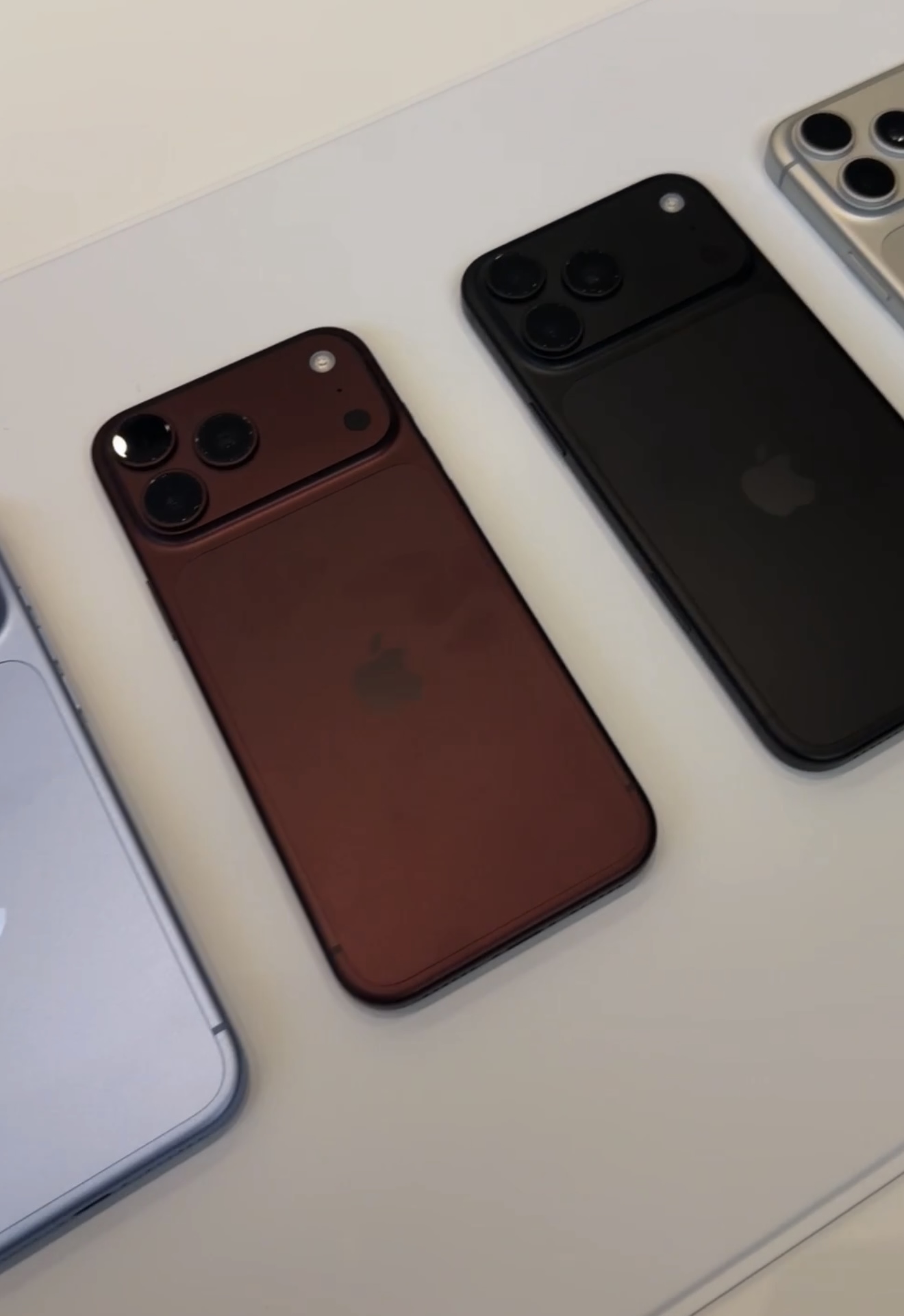 Three iPhone 18 Pro units face down on a white table in Burgundy, Deep Black and Silver, showing the full width camera plateau
