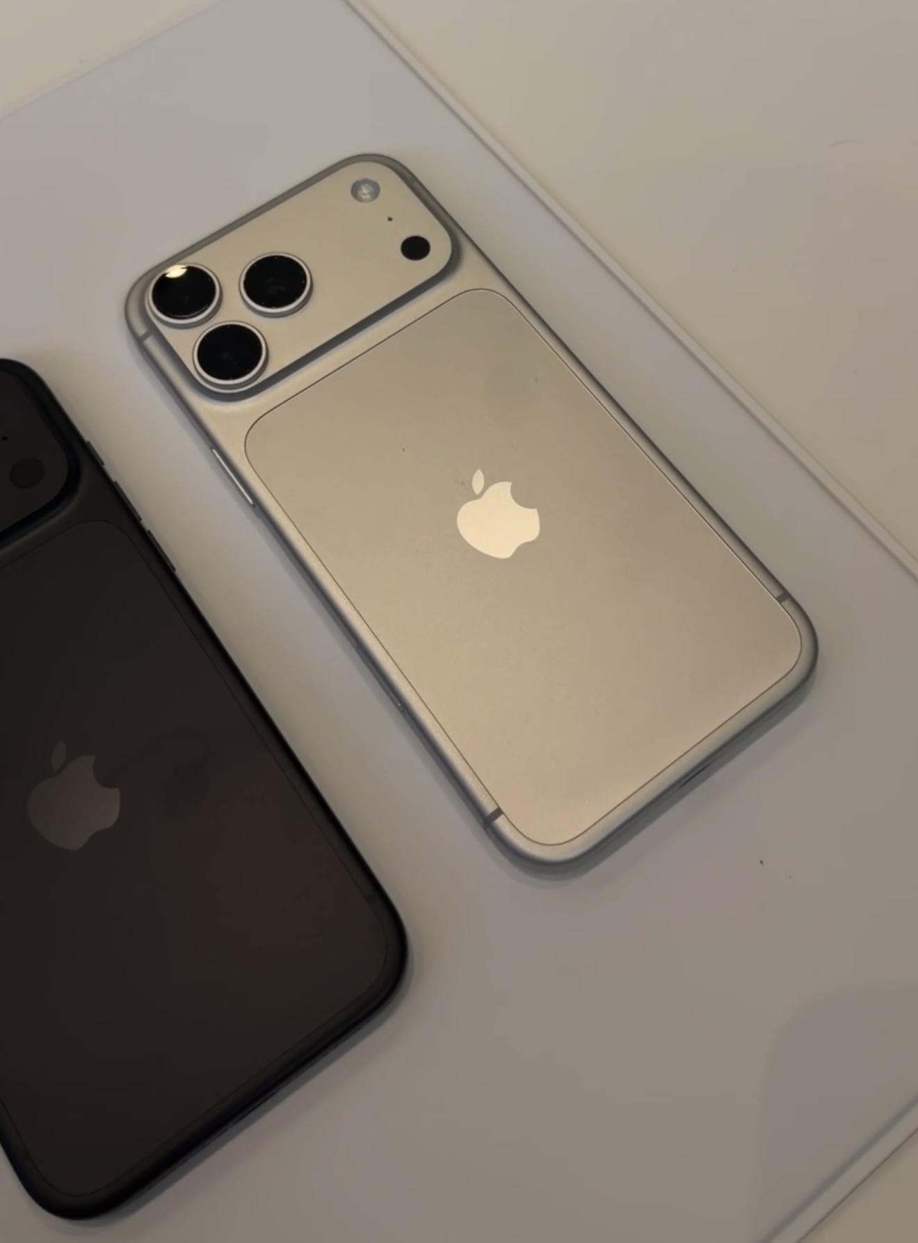 Close up of a Silver iPhone 18 Pro face down beside a Deep Black one, showing the two tone back panel