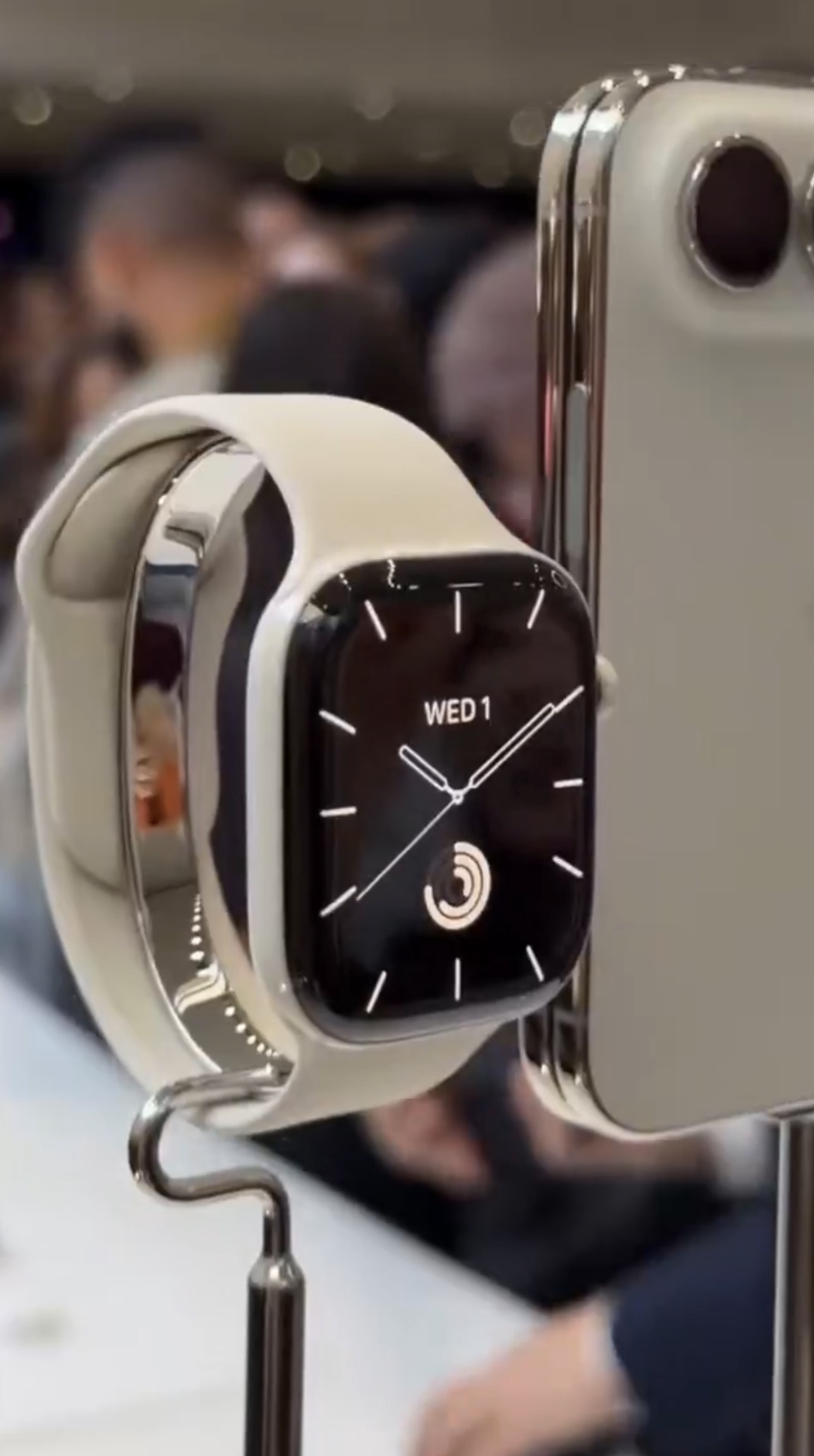 Apple Watch Series 12 on a demo stand with a light sport band, showing an analog watch face reading WED 1