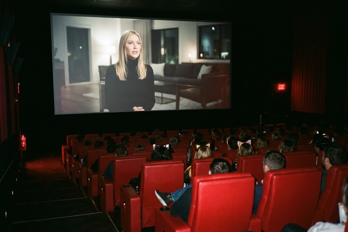 A packed AMC auditorium seen from the back, the screen showing a woman with blonde hair in a black turtleneck in a dimly lit living room, every seat full, a few people in Halloween costumes