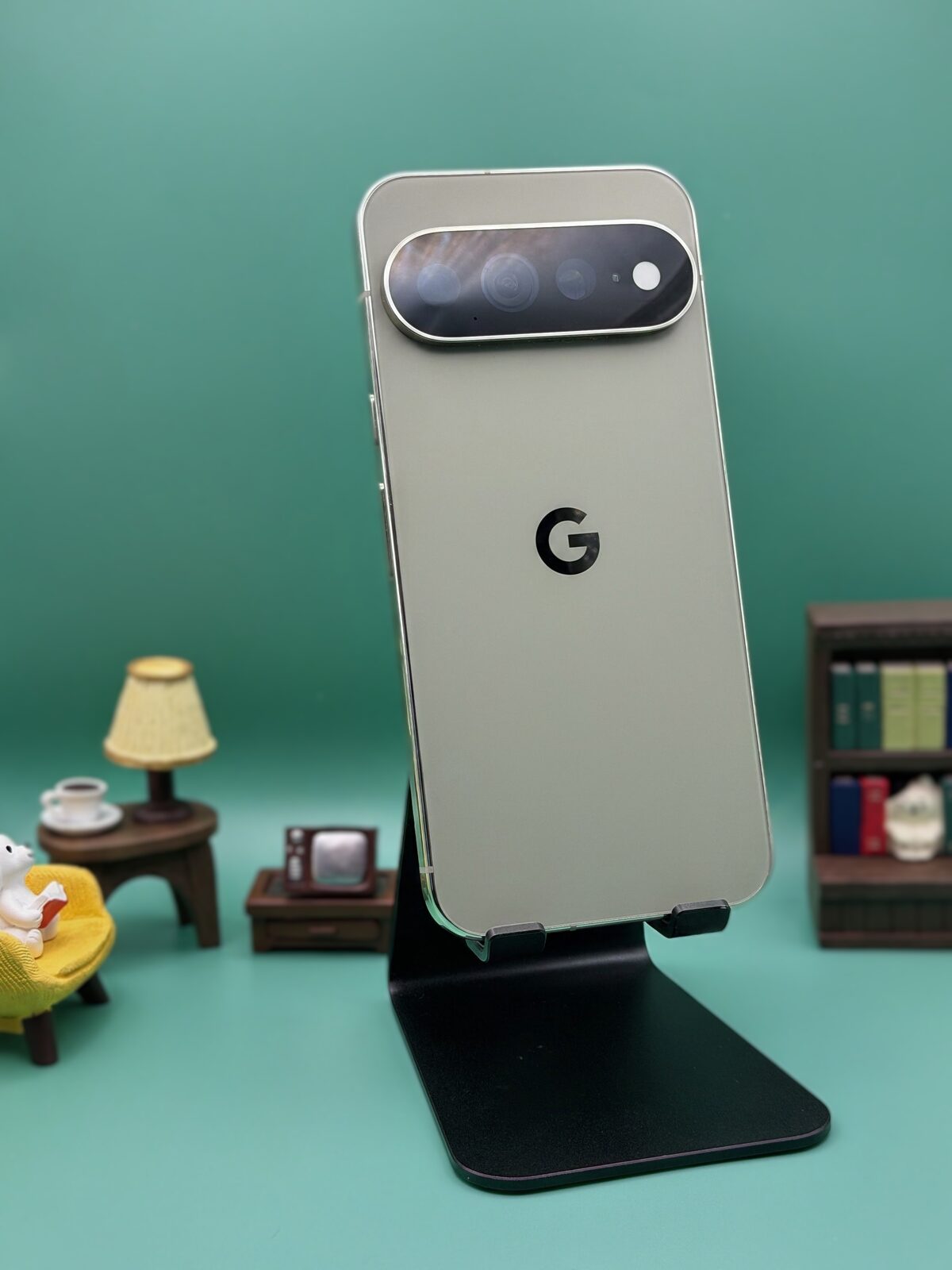 Pixel 11 Pro XL in Olive standing on a black phone stand against a teal background